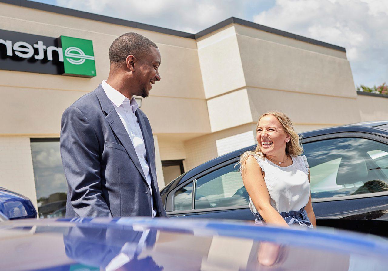 A young man and young woman, both smiling on a sunny day, talk on an Enterprise rental car lot.